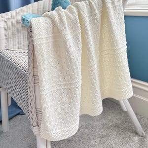 May include: A cream-colored knitted blanket featuring a textured pattern of alternating smooth stockinette sections and rows of small raised bumps. The blanket is draped over a white wicker chair, showcasing its soft, woven construction and edges.