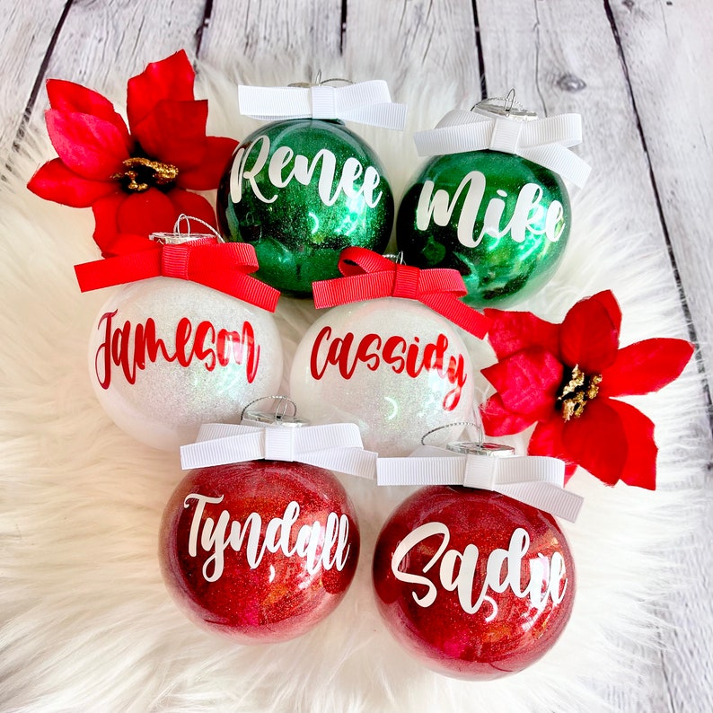 May include: Six personalized Christmas ornaments with glitter. The ornaments are white, green, and red with white bows and red lettering. The names on the ornaments are "Renee", "Mike", "Jameson", "Cassidy", "Tyndall", and "Sadye".