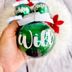 May include: Green glitter Christmas ornament with a white ribbon and the name "Will" written in white cursive lettering.