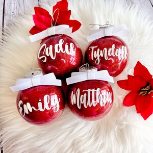 May include: Four red glitter Christmas ornaments with white bows and white lettering. The ornaments are personalized with the names "Sadie", "Tyndall", "Matthew", and "Emily".