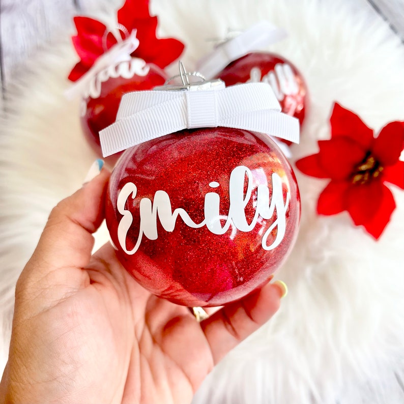 May include: Red glitter Christmas ornament with a white bow and the name "Emily" written in white on the front.