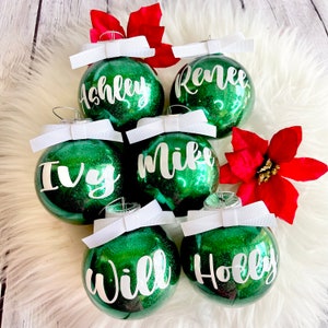 May include: Six green glitter Christmas ornaments with white bows and white lettering. The ornaments are decorated with the names "Ashley", "Renee", "Ivy", "Mike", "Will", and "Holly".