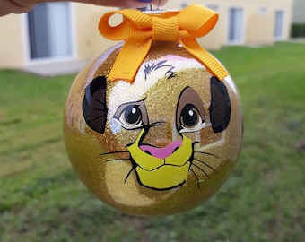Puede incluir: Adorno navideño de purpurina dorada con una imagen de dibujos animados de Simba de El Rey León. El adorno tiene un lazo amarillo.