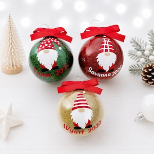 Può includere: Tre palline di Natale rotonde e glitterate in verde, rosso e oro. Ognuna presenta un disegno di uno gnomo con barba bianca, cappello rosso e un fiocco rosso in cima. Le palline sono personalizzate con i nomi Chase, Savanna, Wyatt e l'anno 2022.
