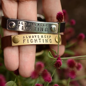 Mosiężna/nierdzewna zawieszka Always Keeps Fighting z czarną lub brązową skórzaną bransoletą ze sprzączką
