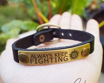 May include: Black leather bracelet with a gold metal plate engraved with the words "Always Keep Fighting" and a symbol.