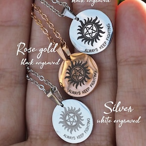 Może przedstawiać: Trzy srebrne naszyjniki z okrągłym wisiorkiem przedstawiającym pentagram w kółku z napisem "Always Keep Fighting". Naszyjniki są pokazane na dłoni, przy czym górny naszyjnik ma grawerowanie czarne, środkowy naszyjnik ma grawerowanie w kolorze różowego złota, a dolny naszyjnik ma grawerowanie białe.