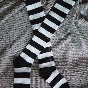 以下が含まれることがあります： 黒と白のストライプのニーハイソックスのペア。靴下は灰色のニットブランケットの上に置かれています。靴下は黒と白のストライプが交互に並んでいます。背景は灰色のニットブランケットです。