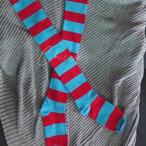 Może przedstawiać: Para skarpetek do kolan z naprzemiennymi czerwonymi i jasnoniebieskimi poziomymi paskami. Skarpetki leżą na szarej dzianinowej narzucie. Skarpetki mają czerwony palec i piętę.