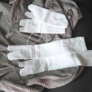 Puede incluir: Calcetines y guantes tabi blancos se muestran sobre un suéter de punto gris. Un collar de cuentas negro está drapeado cerca. Los calcetines y guantes tienen un diseño de punta dividida. La imagen tiene una iluminación suave y natural.