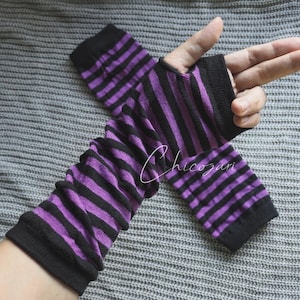 Peut inclure: Paire de mitaines sans doigts à rayures noires et violettes. Les mitaines sont faites d'une matière douce et extensible et présentent un poignet côtelé et un motif à rayures noires et violettes. Les mitaines sont présentées sur un fond gris.