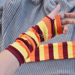 May include: Striped arm warmers in shades of orange, yellow, and burgundy. The arm warmers have a thumb hole and are displayed on a grey background. The text "Chicozari" is visible on the arm warmer.