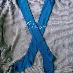 Puede incluir: Un par de calcetines altos azul brillante, cruzados sobre una manta de punto gris. Los calcetines son de color liso, con una textura suave. El texto "Chicozari" está escrito en blanco sobre los calcetines.