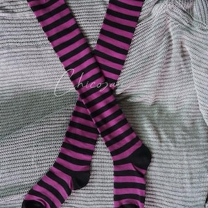 May include: A pair of knee-high socks with alternating black and purple horizontal stripes. The socks have black toes and heels. The socks are lying on a grey knitted fabric.