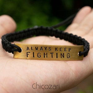 May include: Black braided bracelet with a rectangular gold-colored metal plate. The plate is stamped with the words "ALWAYS KEEP FIGHTING". The bracelet is held in a person's hand.