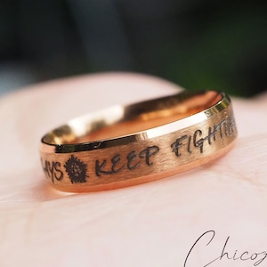 Może przedstawiać: Złoty pierścionek z napisem "Always Keep Fighting" wygrawerowanym na obrączce.