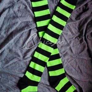 以下が含まれることがあります： 黒とネオングリーンの水平ストライプが交互に配置されたニーハイソックスのペア。靴下は灰色の布の表面に置かれています。「Chicozari」の文字が筆記体で靴下に書かれています。