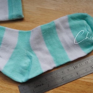 May include: A pair of white and turquoise striped socks. The socks are laid flat on a wooden surface with a ruler next to them.