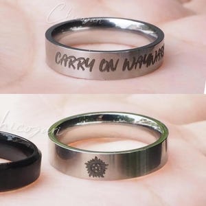 May include: Silver-colored ring with the engraved text "CARRY ON WAYWARD" in a handwritten font. Another silver ring with a sunburst design is also visible. The rings are displayed on a pink surface.