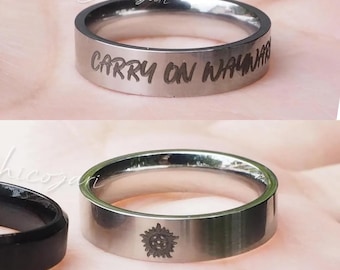 May include: Silver-colored ring with the engraved text "CARRY ON WAYWARD" in a handwritten font. Another silver ring with a sunburst design is also visible. The rings are displayed on a pink surface.
