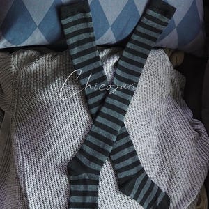 May include: Two knee-high socks with black and grey horizontal stripes. They are laid on a grey knitted jumper. A blue and grey diamond-patterned cushion is in the background.