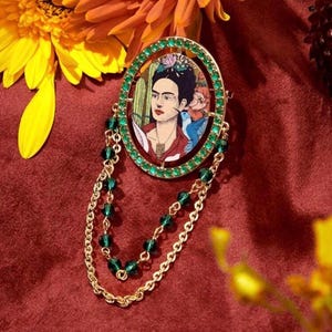 May include: A gold-toned brooch featuring a portrait of Frida Kahlo surrounded by green rhinestones. The brooch is attached to a gold chain with green beads.