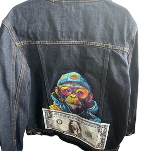 May include: Dark blue denim jacket featuring a vibrant graphic of a monkey wearing sunglasses and a hat, holding a one million dollar bill. The jacket has a classic collar and button closure, with visible stitching.