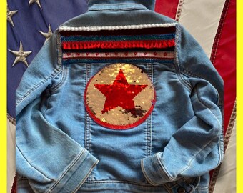 May include: A light blue denim jacket with a red and gold star sequin patch on the back. The jacket has a white, red, and blue trim around the collar and down the back.