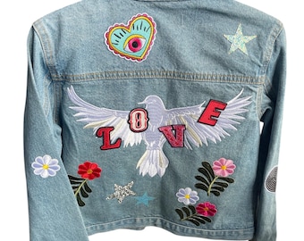 May include: Light blue denim jacket with embroidered patches. Features a dove with "LOVE" lettering, floral and star designs. The jacket has a classic collar and button-up front.