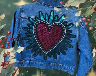 May include: A blue denim jacket with a large sequined heart patch on the back. The heart is red with gold accents and is surrounded by a blue and green sequined design.