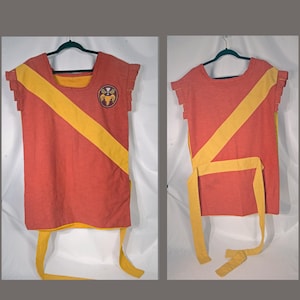 May include: Two-piece costume in shades of red and yellow. The top is a sleeveless tunic with a yellow diagonal stripe and a circular emblem. The bottom is a matching yellow sash. The costume is hanging on a green hanger.