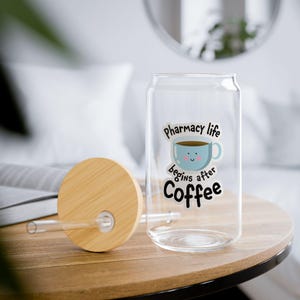 Puede incluir: Un vaso transparente en forma de lata con tapa y pajita de bambú. El vaso tiene una pegatina con una taza de café azul y el texto "Pharmacy life begins after Coffee". La tapa es redonda y de madera clara.