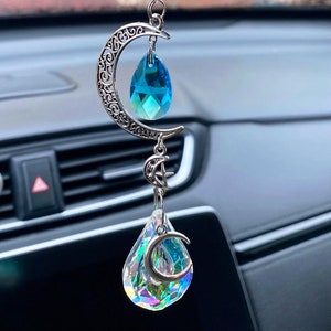 May include: A silver crescent moon charm with a blue crystal teardrop hanging from it. The charm is attached to a chain with a silver clip and a string of blue and clear crystal beads.