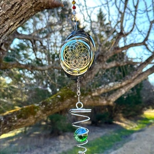 May include: A silver wind chime with a spiral design and a green glass bead. The chime is hanging from a tree branch and has a rainbow-colored beaded chain.