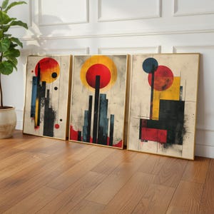 May include: Three framed abstract art prints. Each print features a gold frame and a unique design with red, yellow, blue, and black geometric shapes. The artwork is displayed on a light-colored background with a wood floor.