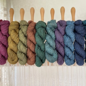 May include: Eight skeins of yarn in various shades of green, blue, and purple are displayed on a wooden loom. The yarn is arranged in a rainbow pattern, with the darkest shades at one end and the lightest shades at the other.