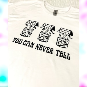 May include: White t-shirt with a black graphic of three wells with buckets and the text "YOU CAN NEVER TELL" printed in a retro font.