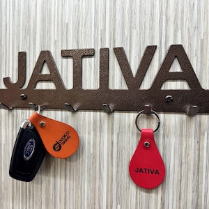 May include: A metal key rack with the word "JATIVA" in large letters. The rack has five hooks and two keychains hanging from it. One keychain is orange with the text "DECOR SIGNS" and the other is red with the text "JATIVA".