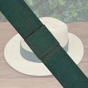 May include: A dark green hat band is draped over a cream-colored Panama hat. The hat band is made of a textured material and is secured with a black band. The hat is resting on a wooden surface.