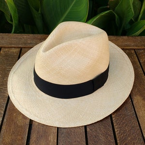 May include: A light-colored Panama hat with a black band. The hat is made of woven straw and has a classic fedora shape. The hat is sitting on a wooden surface.