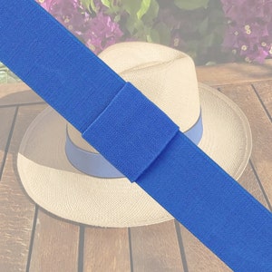 May include: A bright blue hat band is displayed diagonally. The band, made of textured fabric, contrasts with a straw hat featuring a blue ribbon. The hat rests on a wooden surface, creating a summery feel.