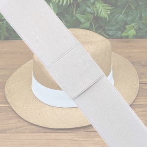 May include: A white elastic band with a woven texture. The elastic band is lying on a wooden surface with a straw hat in the background.