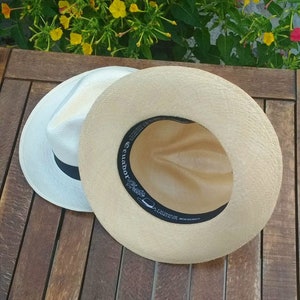 May include: Two Panama hats, one white with a black band and the other natural beige, sit on a wooden surface. The interior of the beige hat displays a black band with white text that reads "Ecuador Hats".