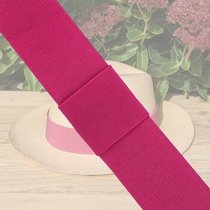 May include: A straw hat with a pink ribbon and a wide magenta band. The hat is set against a background of flowers and a light wood surface. The magenta band is approximately 7.6 cm wide.
