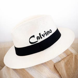 May include: A white fedora hat with a black band and the word "Calvino" printed in black. The hat is made of woven material and has a classic, stylish design. The hat is a summer accessory.
