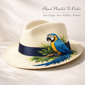May include: A cream-coloured fedora hat with a navy blue band. The hat features a hand-painted blue and yellow macaw parrot design with green leaves. Text on the image reads: Hand Painted To Order. Your Design • Pets • Wildlife • Flowers.