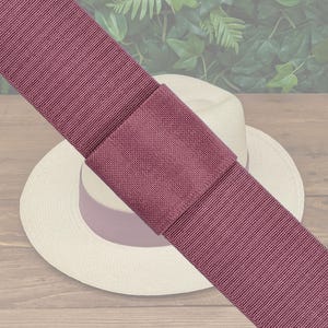 May include: A wide, mauve ribbon with a textured weave, folded over itself and placed on top of a white straw hat. The hat is sitting on a wooden surface.