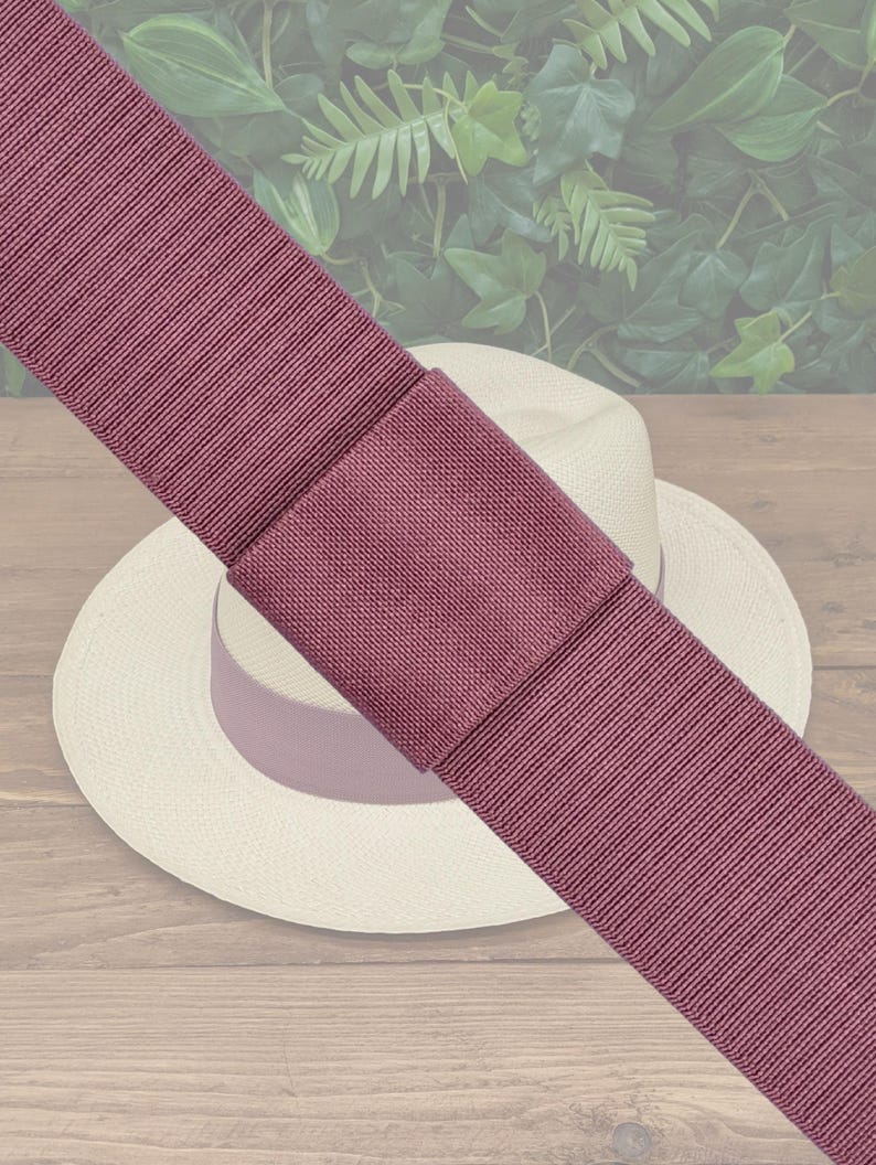 May include: A wide, mauve ribbon with a textured weave, folded over itself and placed on top of a white straw hat. The hat is sitting on a wooden surface.