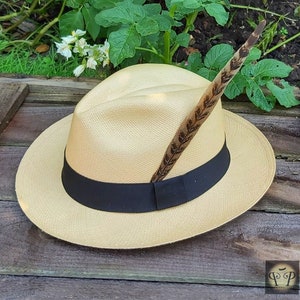 May include: A light tan fedora hat with a black band and a decorative feather. The hat is made of woven material and sits on a wooden surface. The feather is brown with dark brown markings.