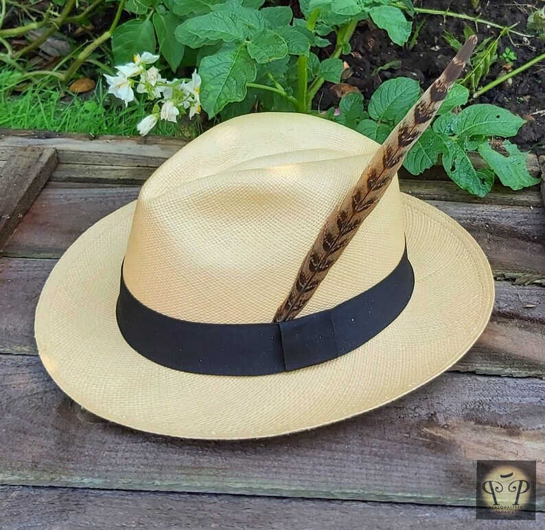 May include: A light tan fedora hat with a black band and a decorative feather. The hat is made of woven material and sits on a wooden surface. The feather is brown with dark brown markings.
